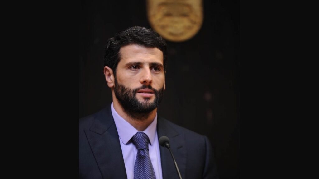Aleksandar Šapić govori za mikrofonom, u tamnom odelu, ispred grba.