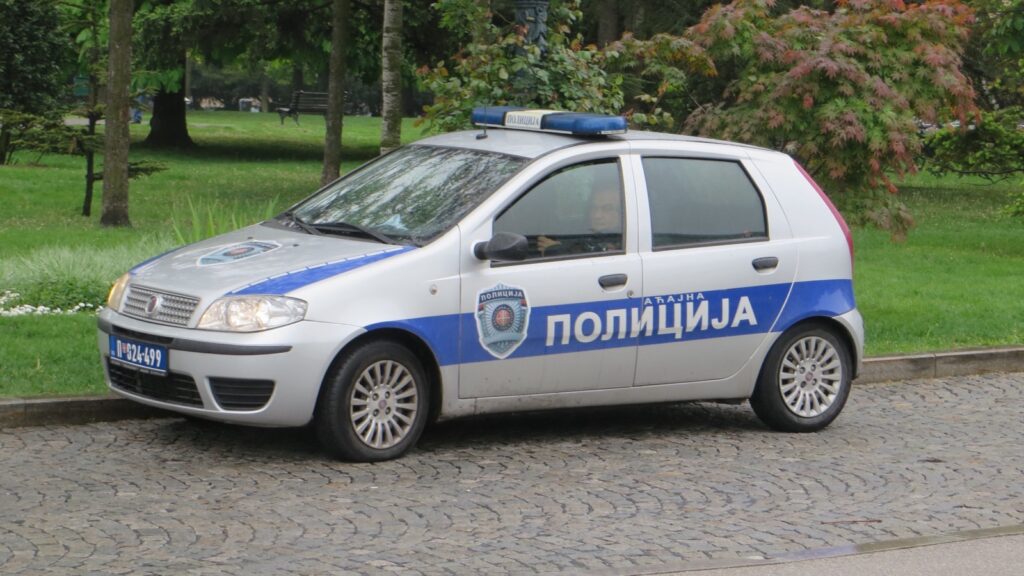 Srebrni policijski automobil sa plavom trakom i natpisom Policija parkiran na kaldrmi pored parka