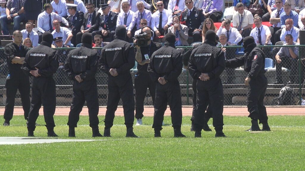 Pripadnici Žandarmerije u crnim uniformama i fantomkama stoje leđima okrenuti kameri ispred tribine sa zvanicama