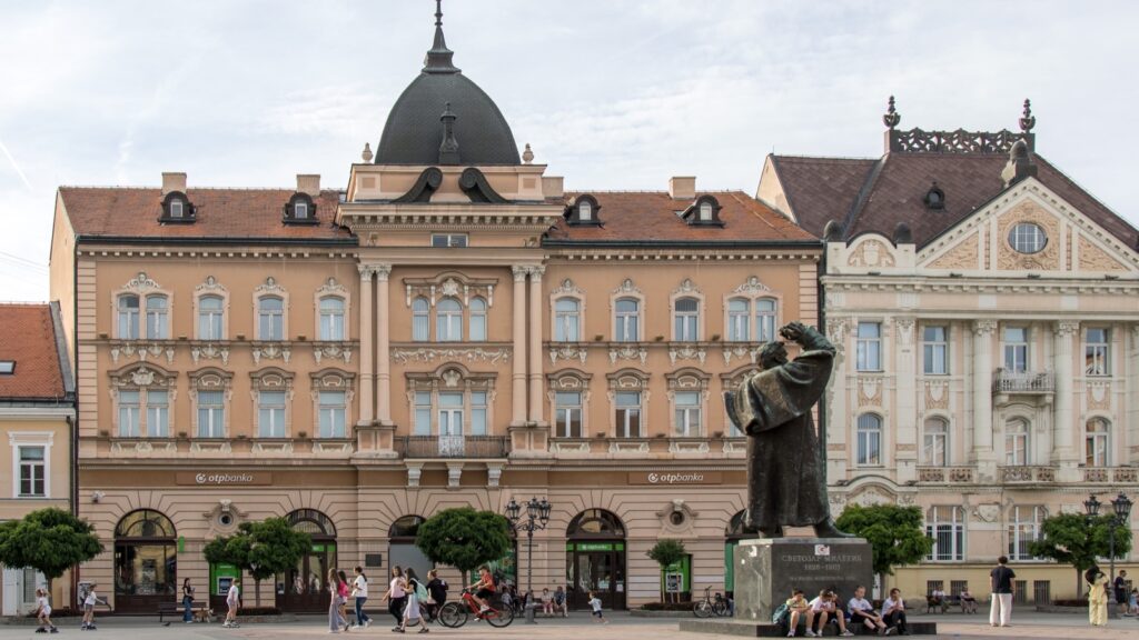 Trg slobode u Novom Sadu sa spomenikom Svetozaru Miletiću ispred zgrade sa kupolom, prolaznici na trgu
